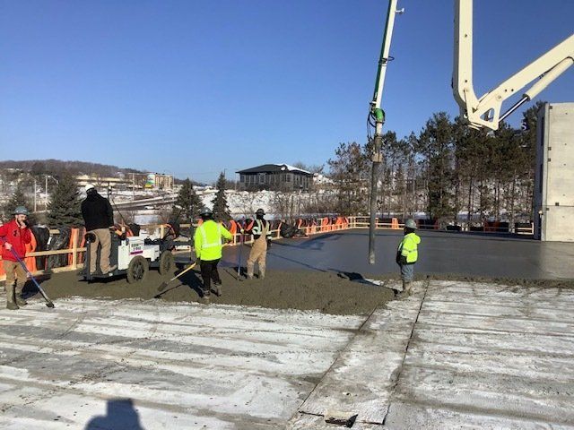 Construction workers pouring concrete on a flat, snow-covered surface with a pump truck under a clear, sunny sky.