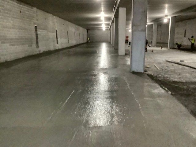 Newly poured, wet concrete floor in an unfinished, dimly lit interior. Workers are visible in the background.