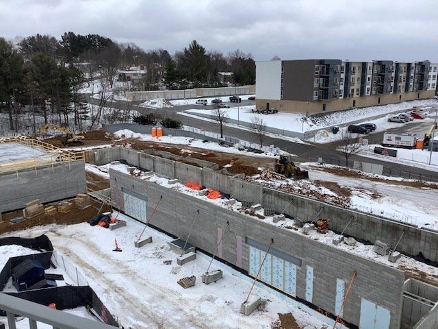Construction site with concrete walls and snow, adjacent to a road and multi-story apartment building under a cloudy sky.