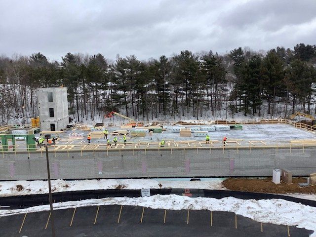 Construction site: Workers on a concrete foundation, building framing with equipment. Winter setting, snow and trees in background.
