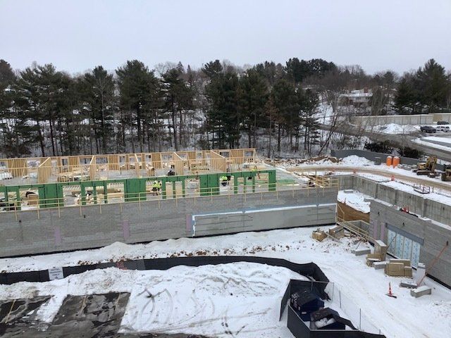 Construction site in winter; wooden frames atop concrete foundation; snow on the ground; trees in background.