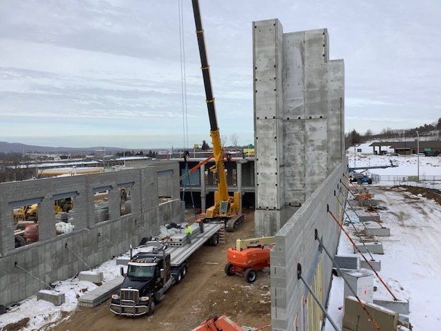 Construction site with crane lifting concrete panels. Gray walls, truck, orange lift, snowy ground.