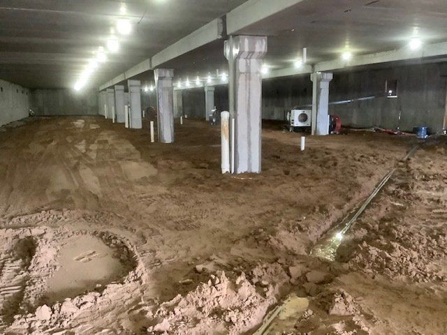 Under-construction space under a bridge with dirt floor, support columns, overhead lights, and exposed plumbing.