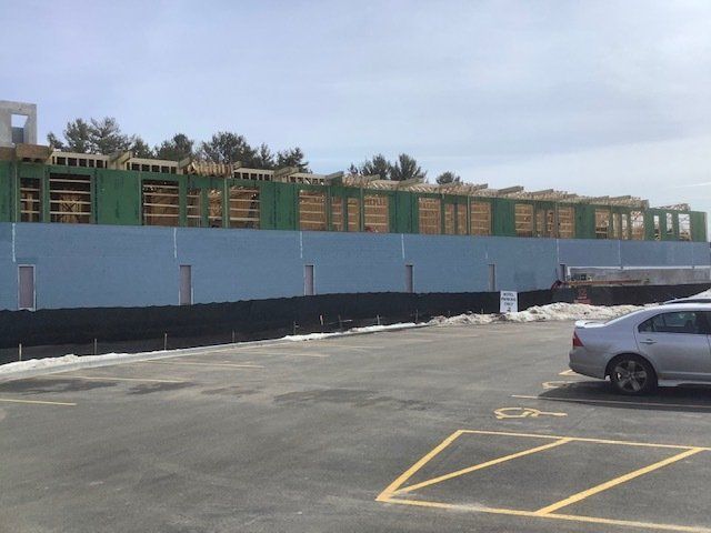 Construction site with blue siding and exposed wood framing. Gray car in parking lot.