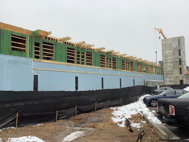 Construction site with building frame, insulation, and snow. Cars parked nearby. Cloudy sky.