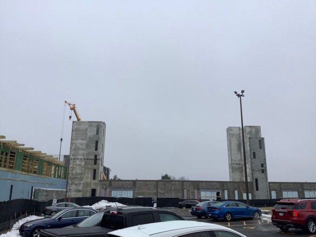 Construction site with two tall concrete towers, crane, and parked cars under an overcast sky.