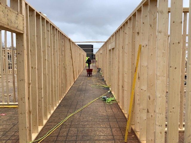 Construction site with wooden wall frames, concrete floor, person with wheelbarrow, cloudy sky.
