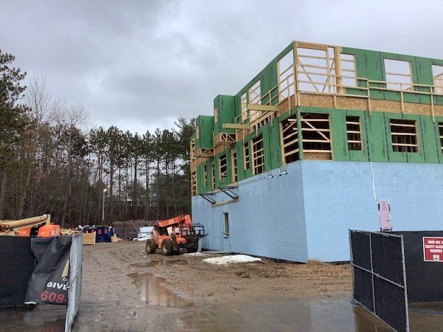 Construction site with a partially built two-story building. Walls are blue; upper levels are wood-framed. Cloudy day.