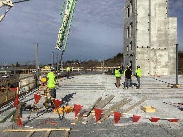 Construction site with workers, concrete pumping, and a large concrete structure under a cloudy sky.