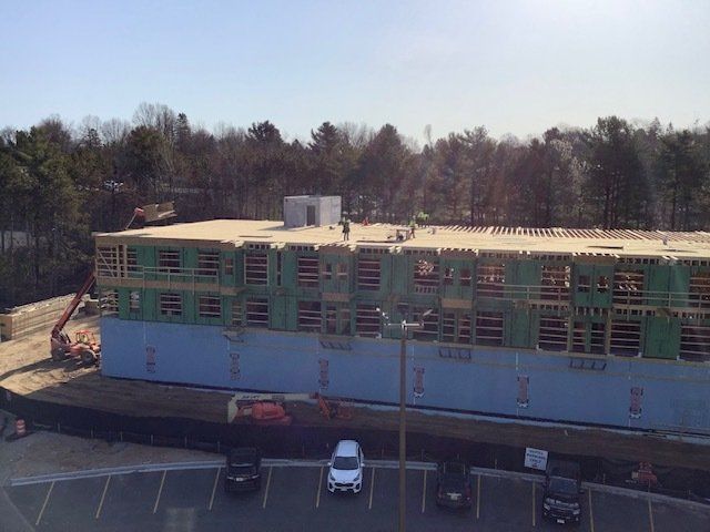 Construction site of a multi-story building, with workers on the roof, visible parking lot in front, and trees in the background.