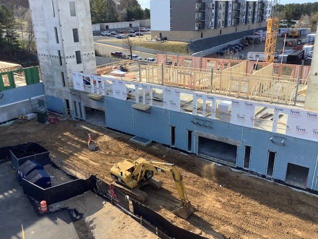 Construction site: excavator on dirt, unfinished building with blue siding, wood framing, and tower.