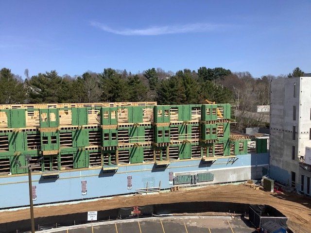 Construction site: wood-framed townhouses under construction, blue foundation, next to a concrete building, sunny day.
