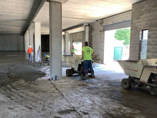 Construction workers pouring concrete on a floor inside a building. Gray concrete, cement pillars, and two motorized wheelbarrows.