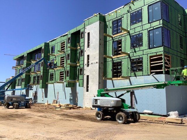 Construction site: multi-story building with workers, lifts, green sheathing, and large windows.