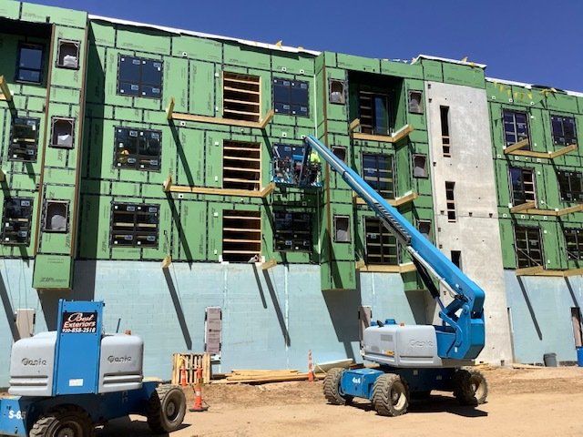 Construction worker in a blue lift working on building exterior; green sheathing, dark windows, and blue sky.