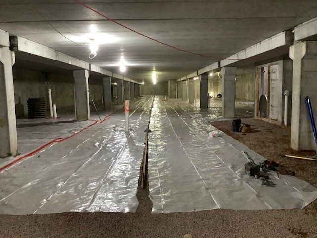 Underground construction site with plastic sheeting covering the ground, concrete pillars, and overhead lighting.