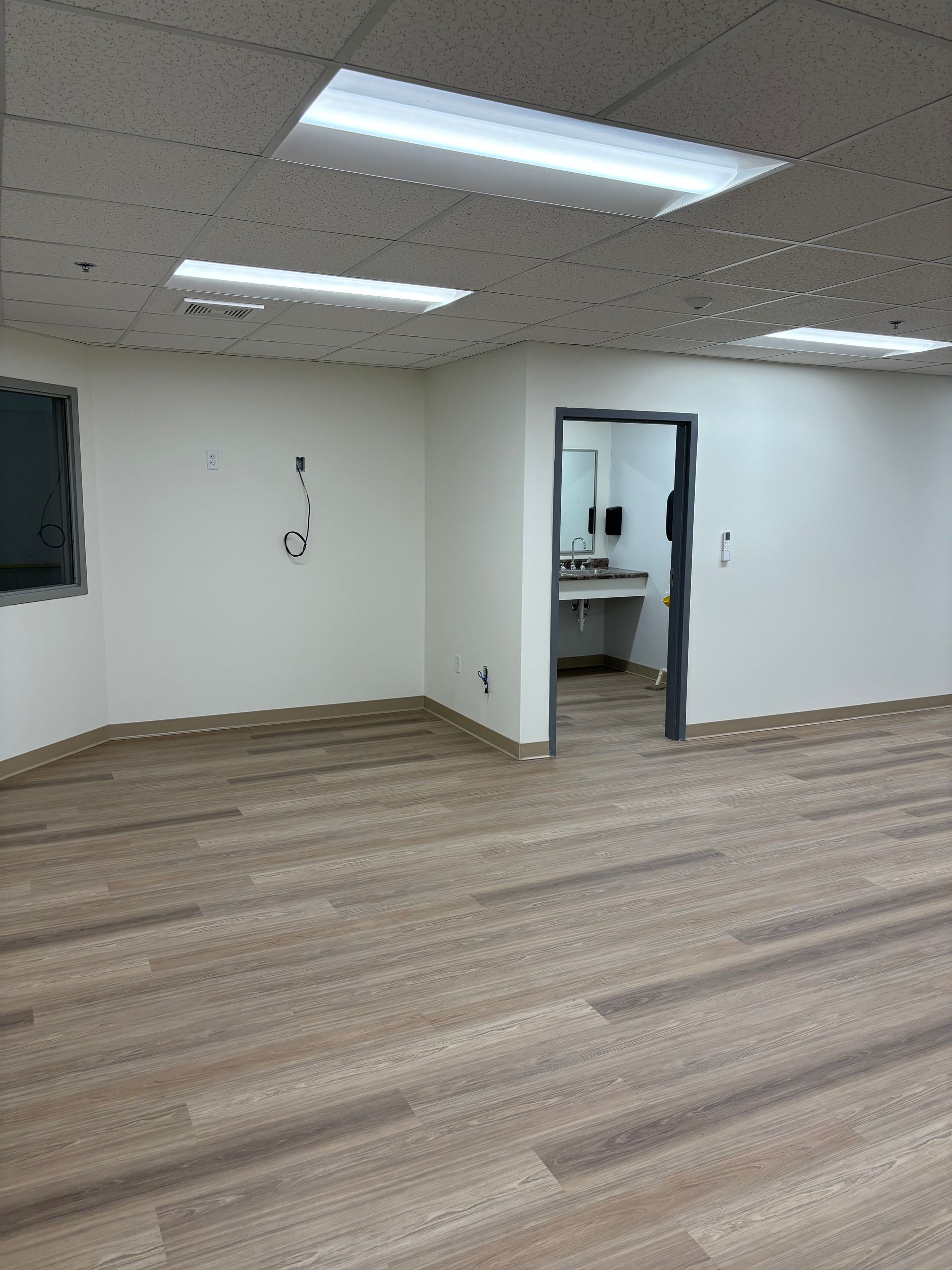 Empty room with neutral walls, wood-look flooring, and a doorway to a restroom. Fluorescent lights illuminate the ceiling.