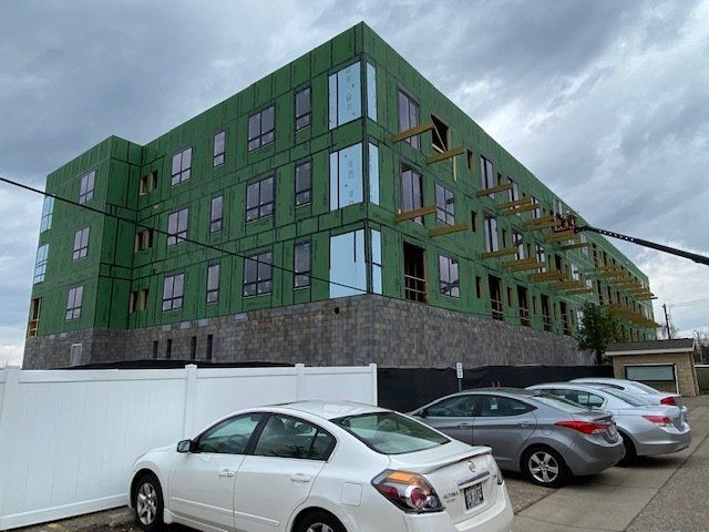 Multi-story building under construction with green exterior, windows, parked cars. Cloudy sky.