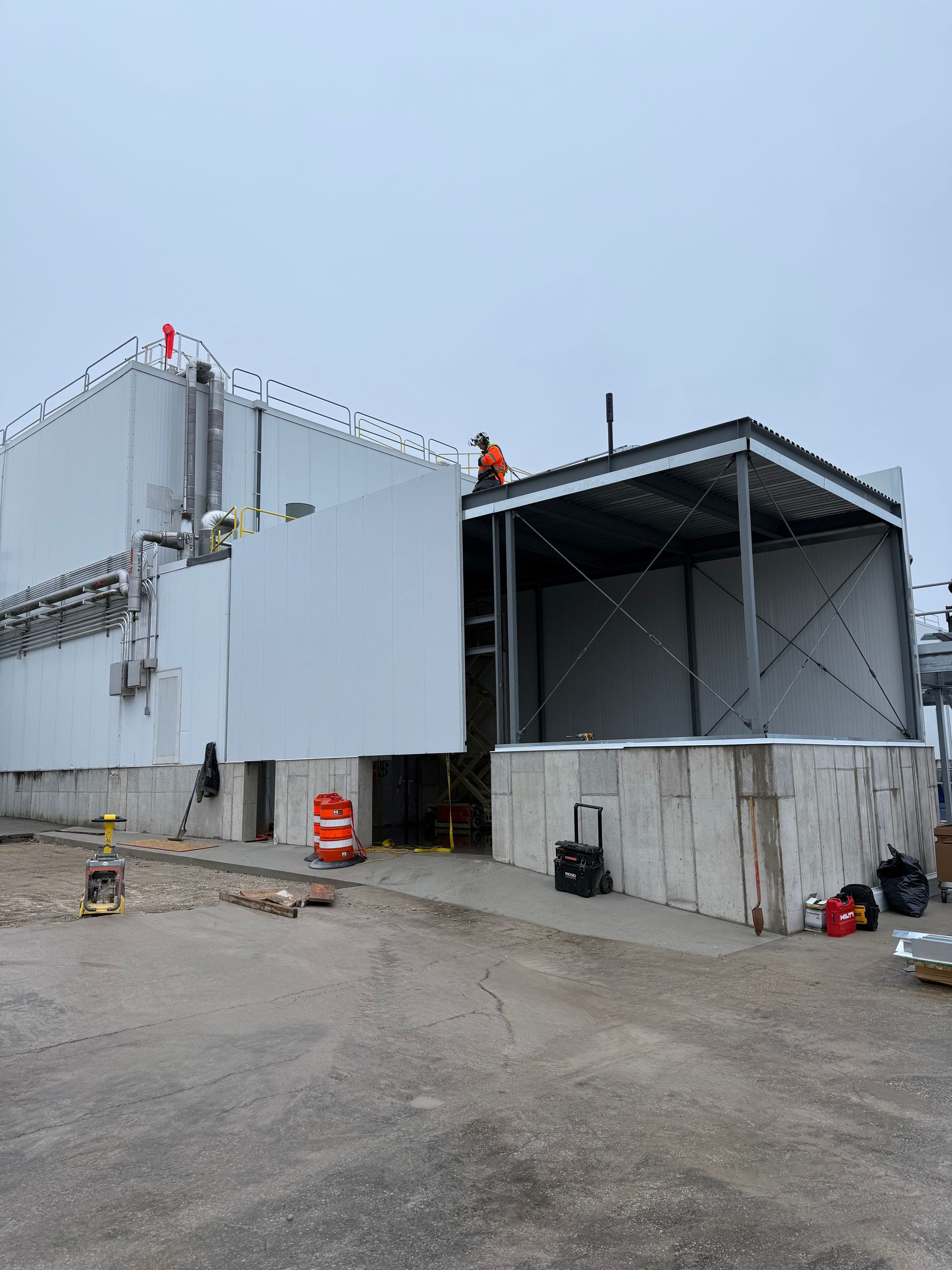 Construction site: Workers on a large, light grey industrial building with a steel frame structure being built to the side. Overcast sky.