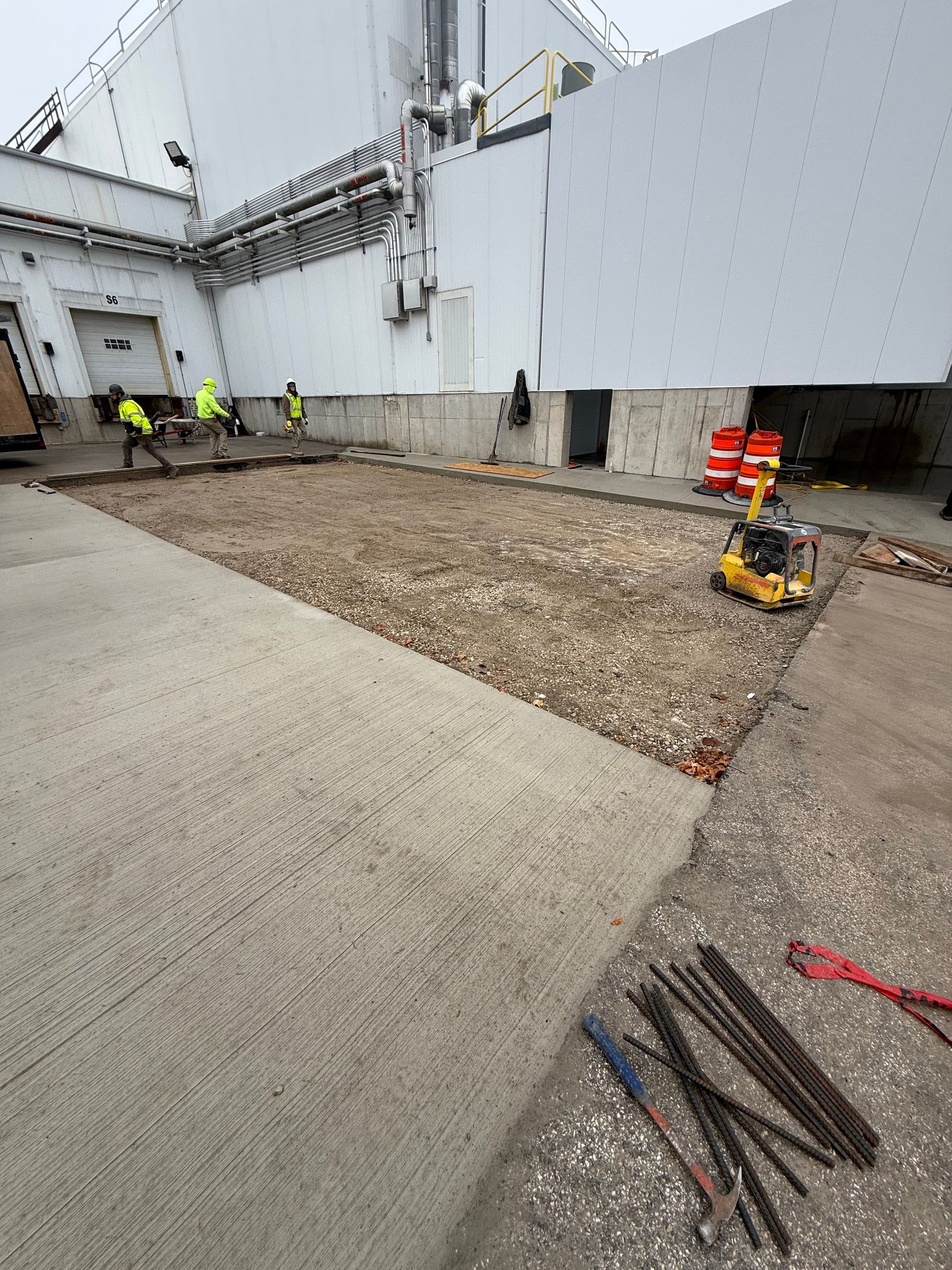 Construction workers preparing a concrete pad near a white building; tools and compacting machine visible.