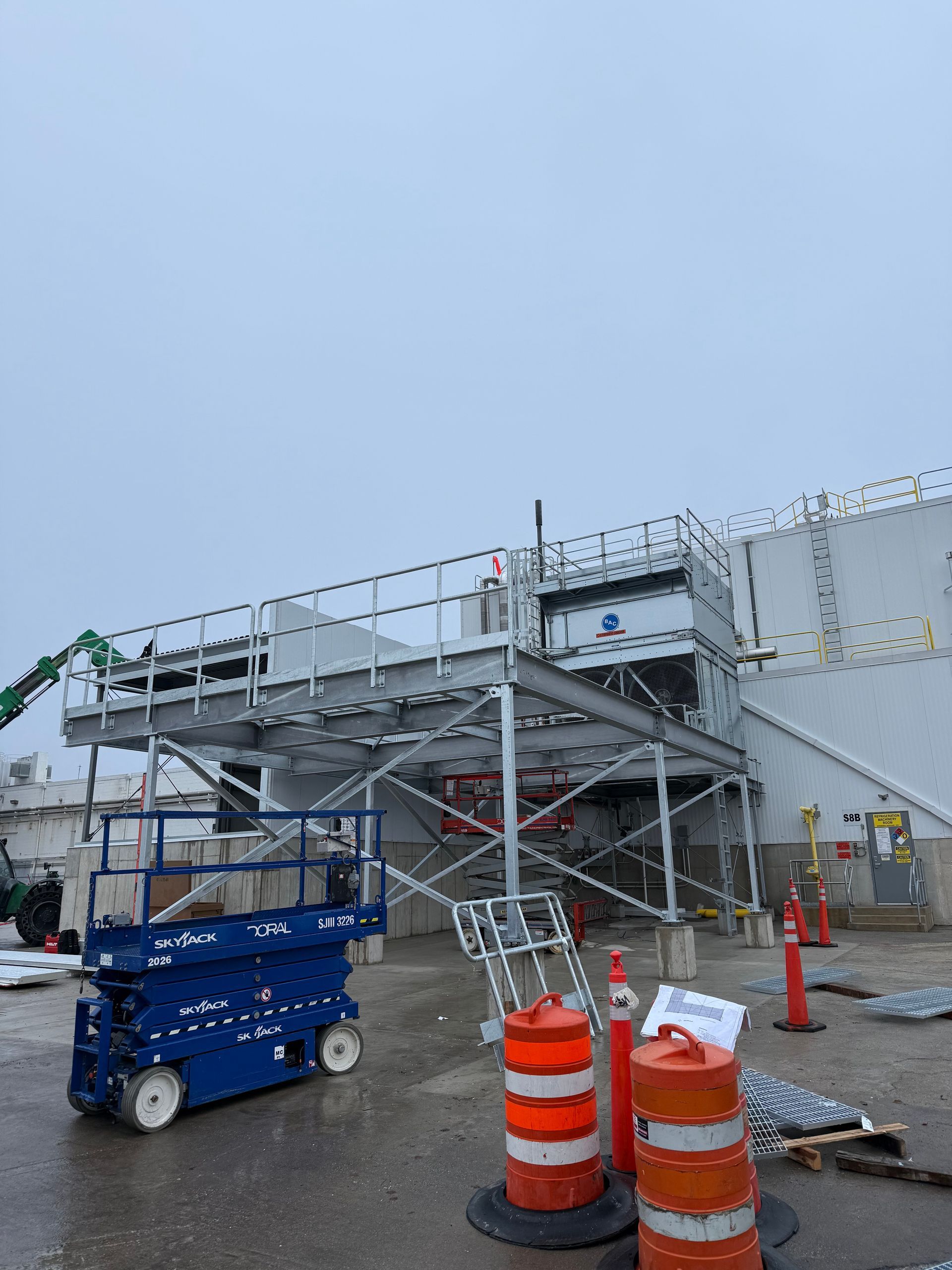 Exterior view of a building with a metal scaffolding structure and a blue lift. Orange traffic cones are present.