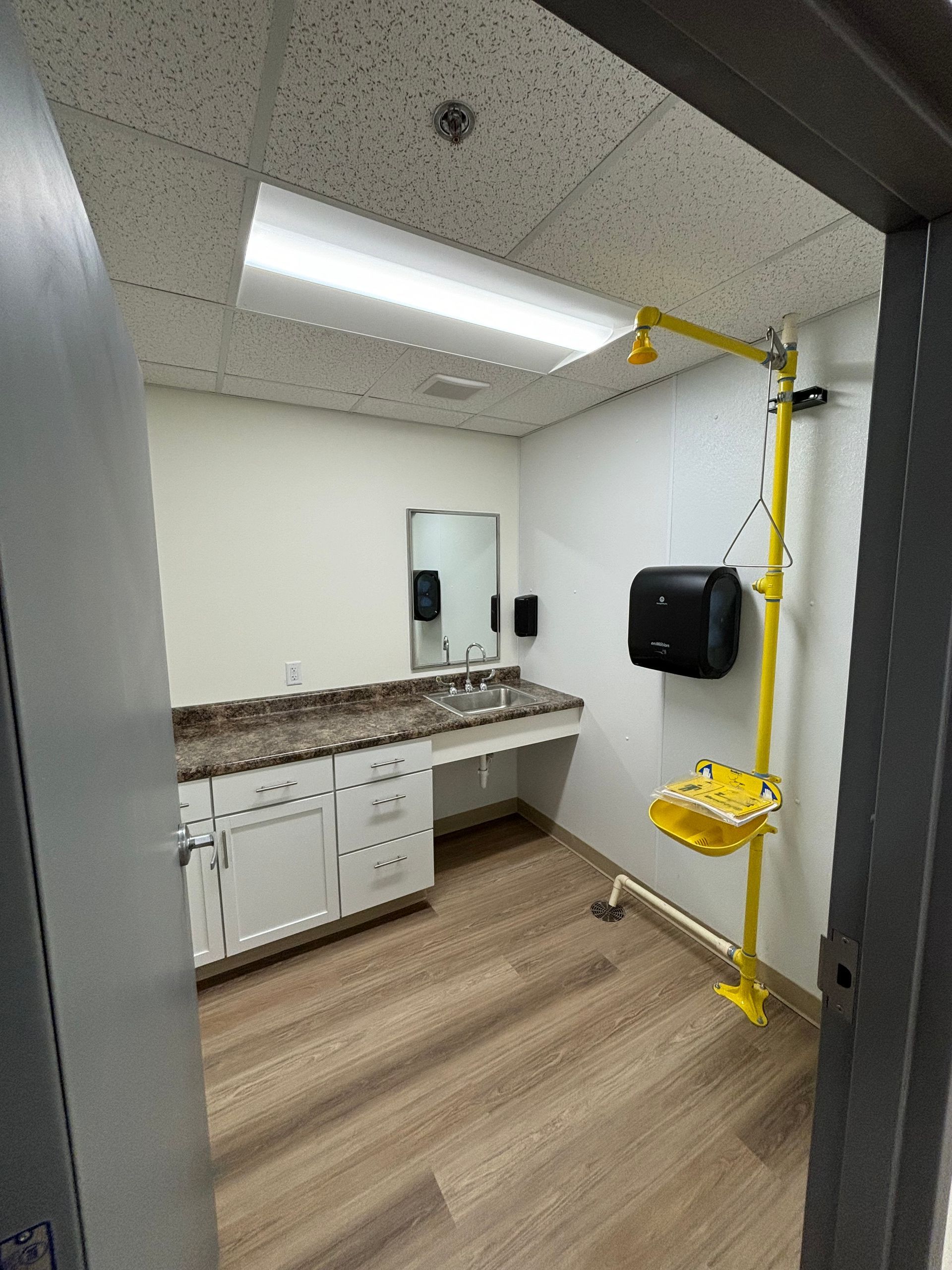Yellow emergency shower in a well-lit room with sink, mirror, and cabinet.