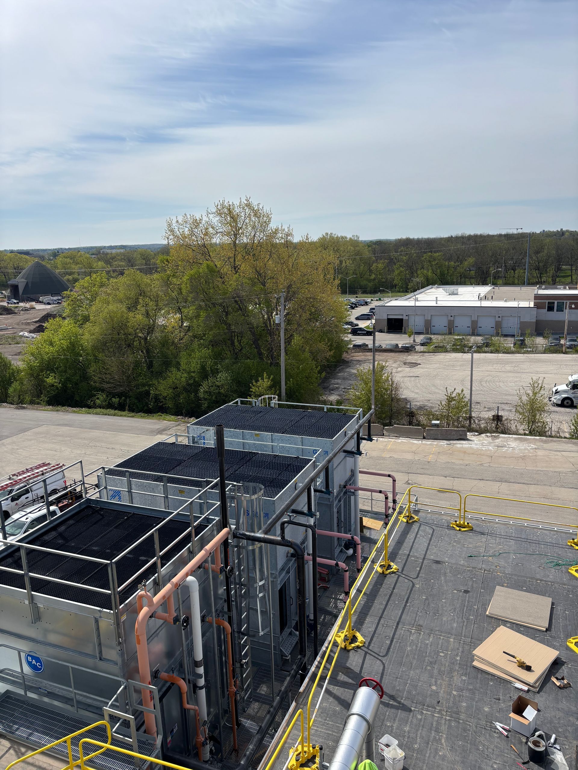 Industrial rooftop with HVAC units and construction materials overlooking trees and low buildings under a cloudy sky