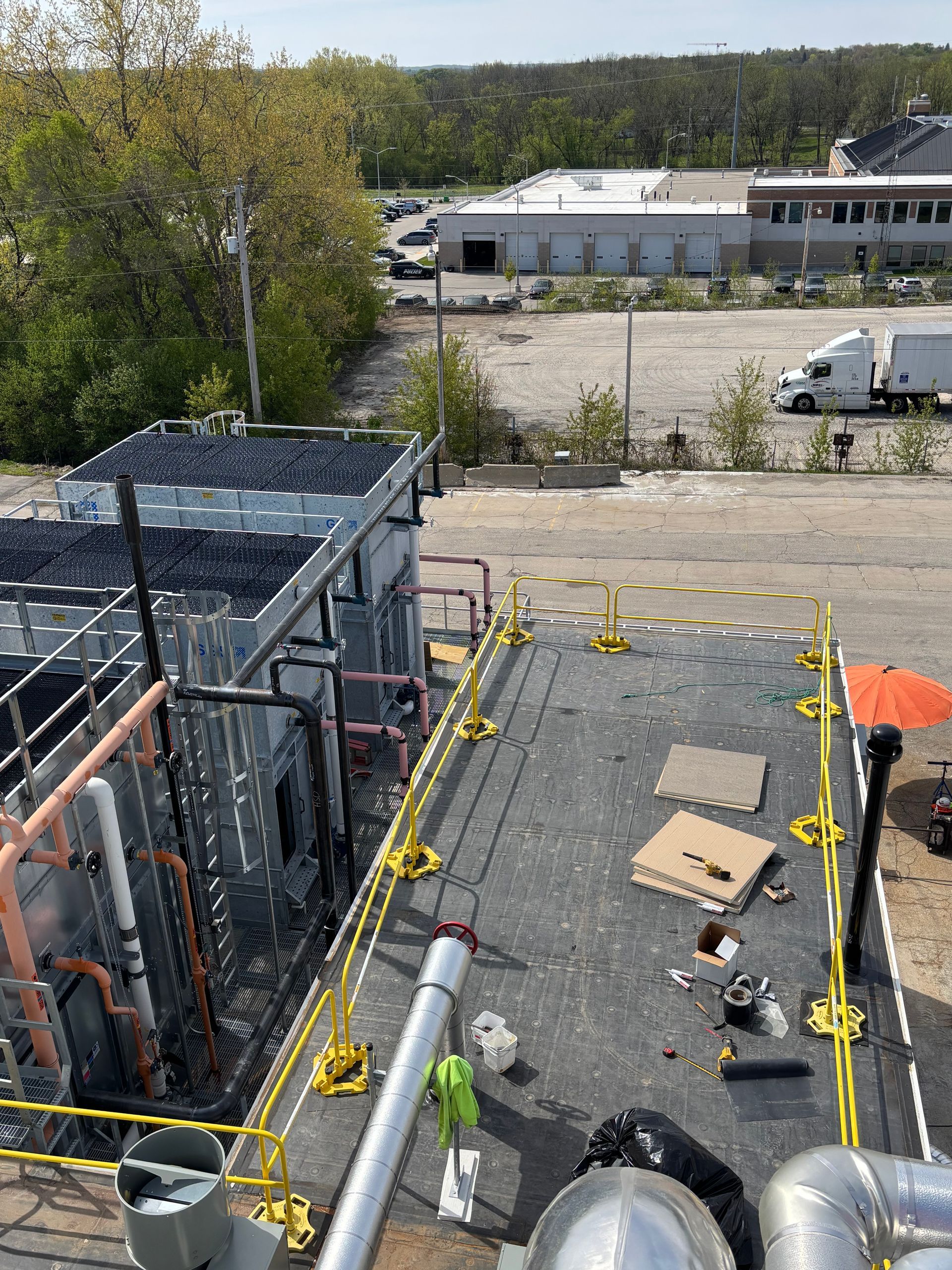 Rooftop construction area with safety rails, tools, cardboard, and a worker in an orange shirt near a parking lot.