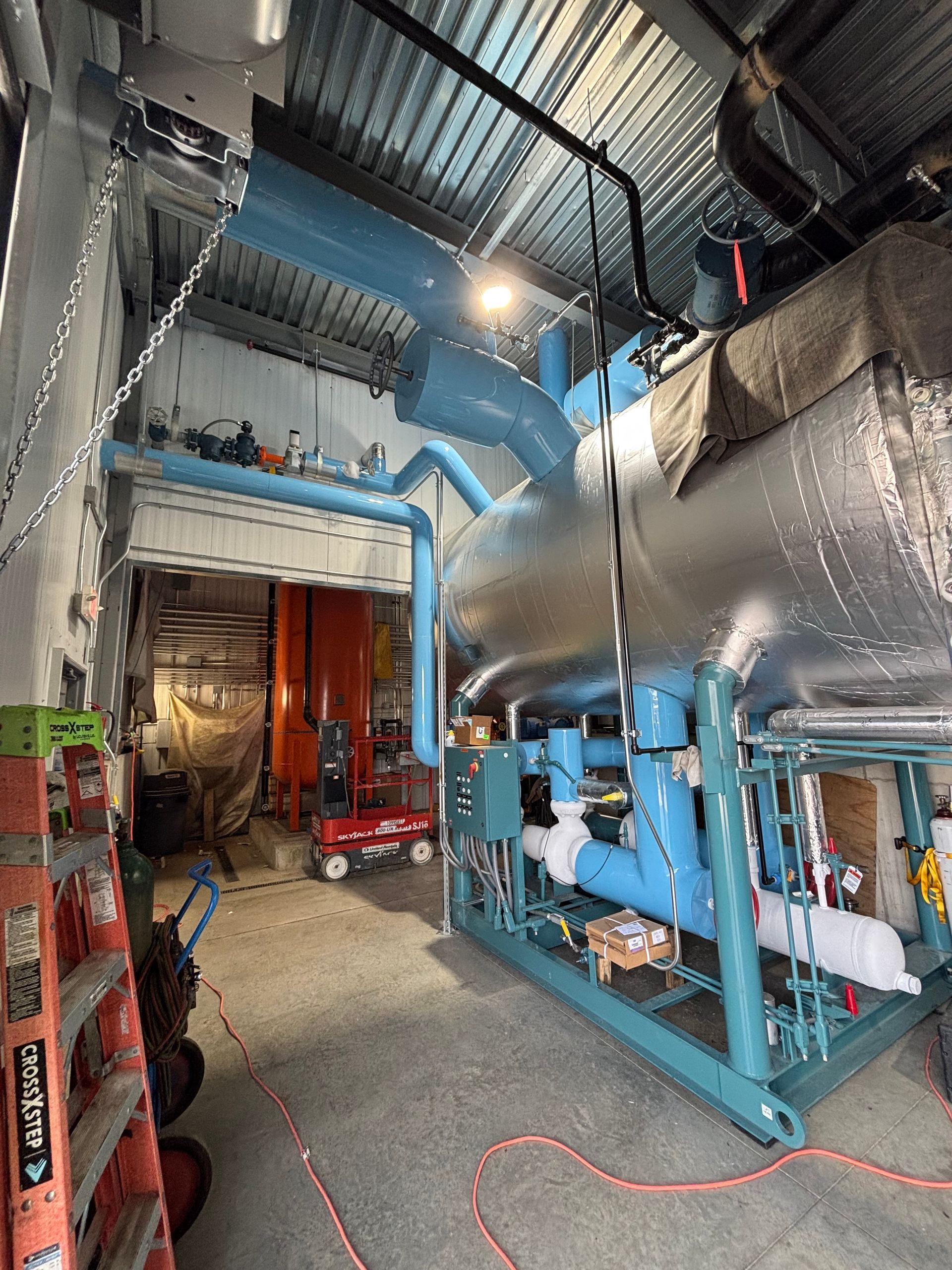 Industrial equipment room with blue pipes, silver tanks, and red machinery under dim overhead lights