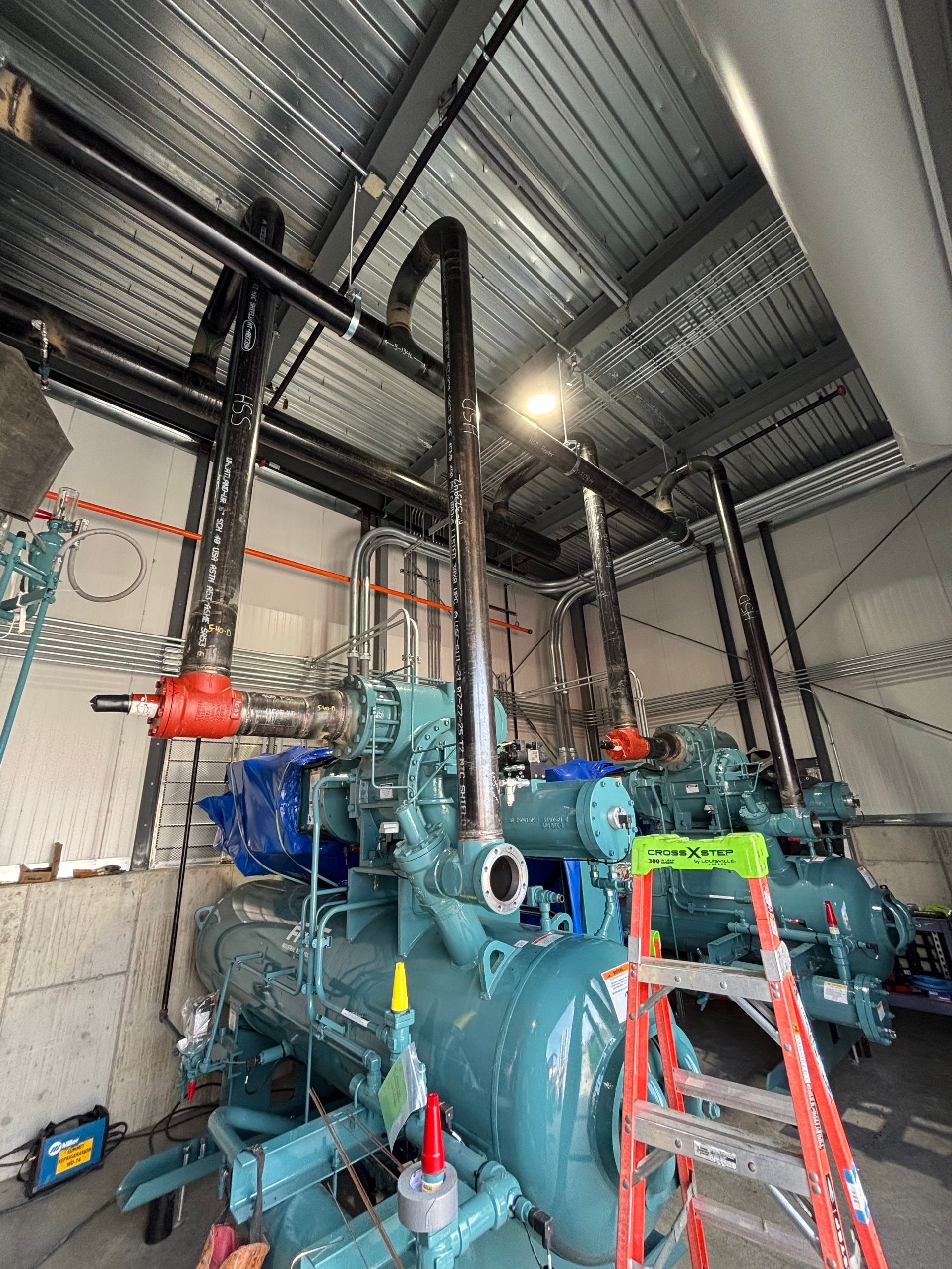 Industrial pump room with a large blue machine, red ladder, and exposed ceiling pipes