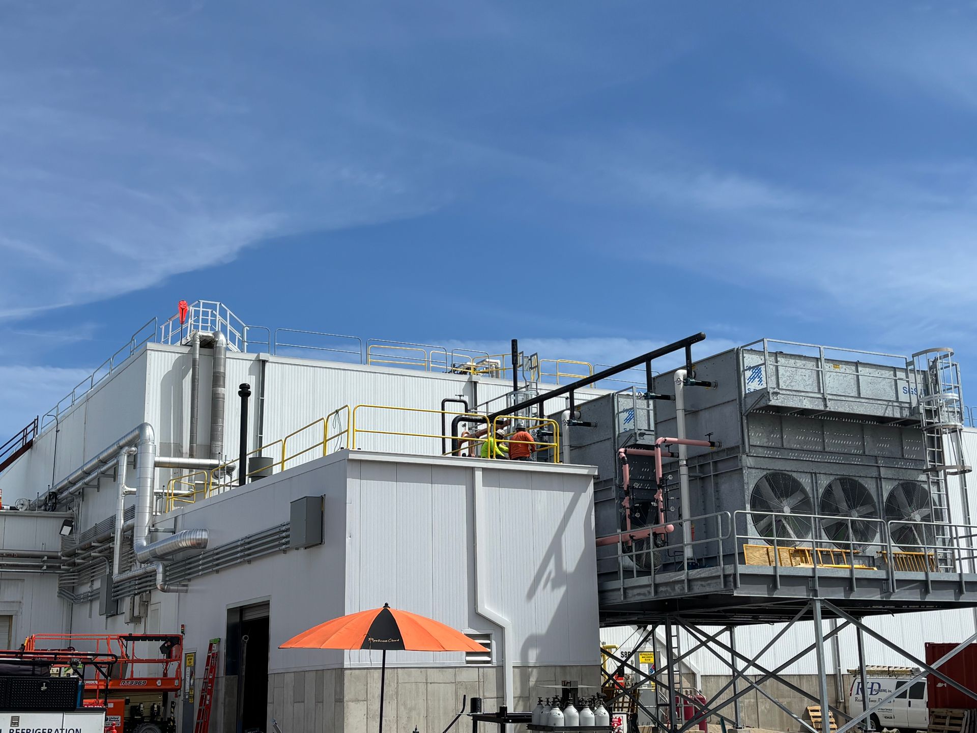 Industrial facility with large metal structures and scaffolding under a blue sky, with an orange umbrella in front.