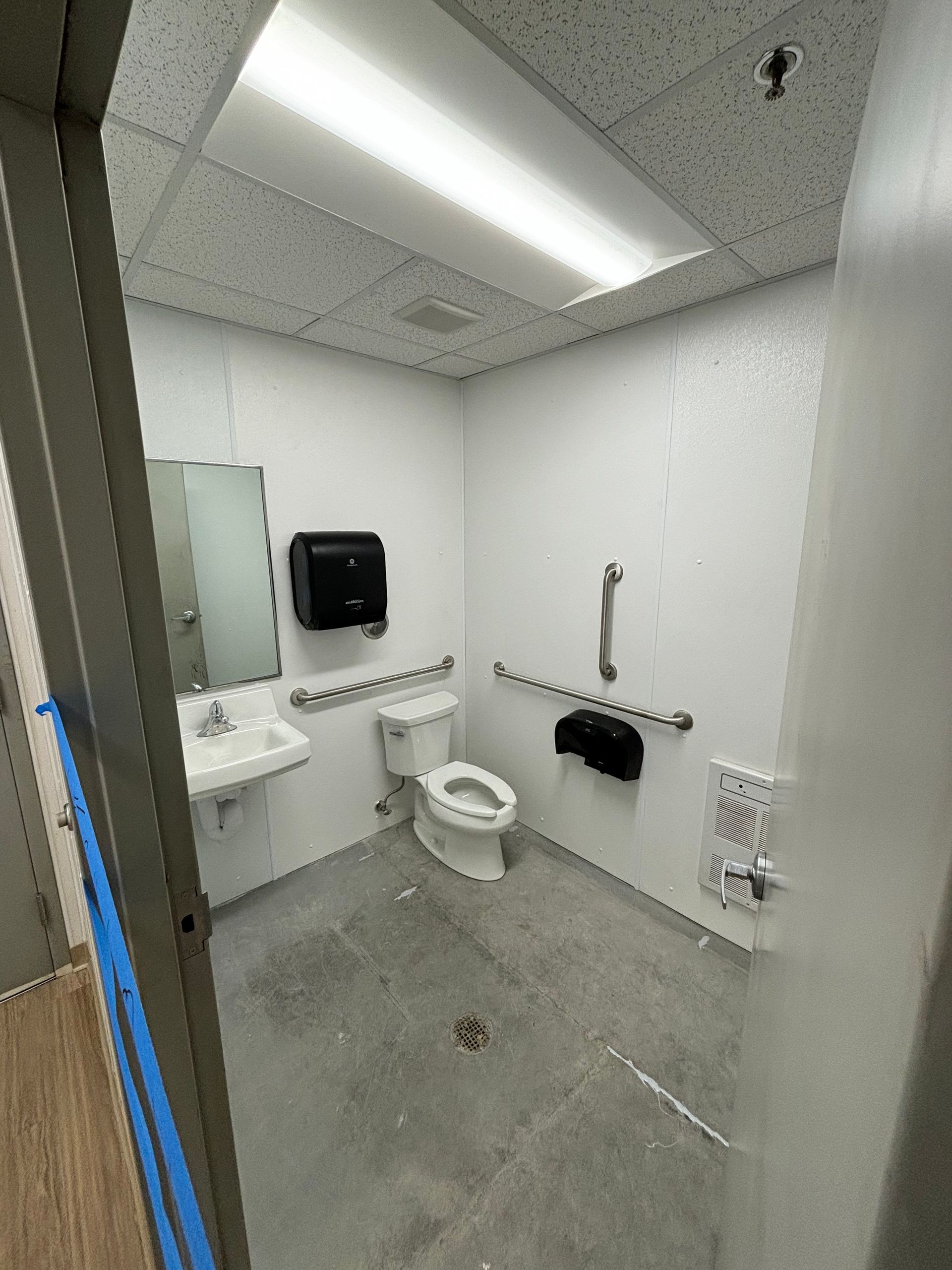 Accessible restroom with toilet, sink, grab bars, and paper towel dispenser. White walls, gray floor.