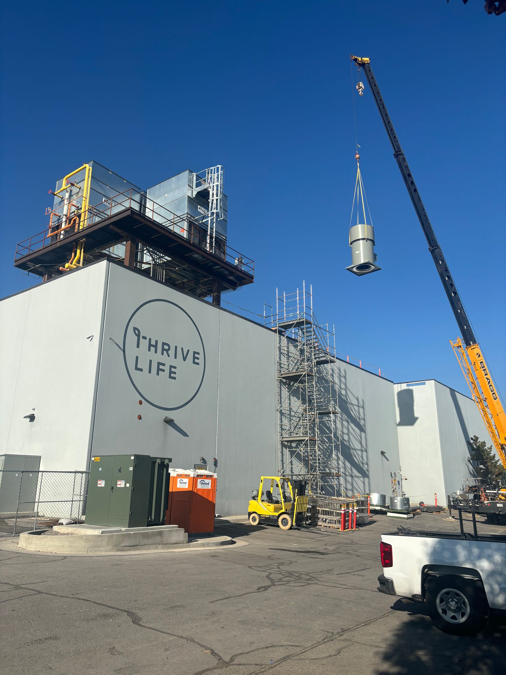 A crane lifting a silver cylindrical object near a large Thrive Life building under construction on a sunny day.