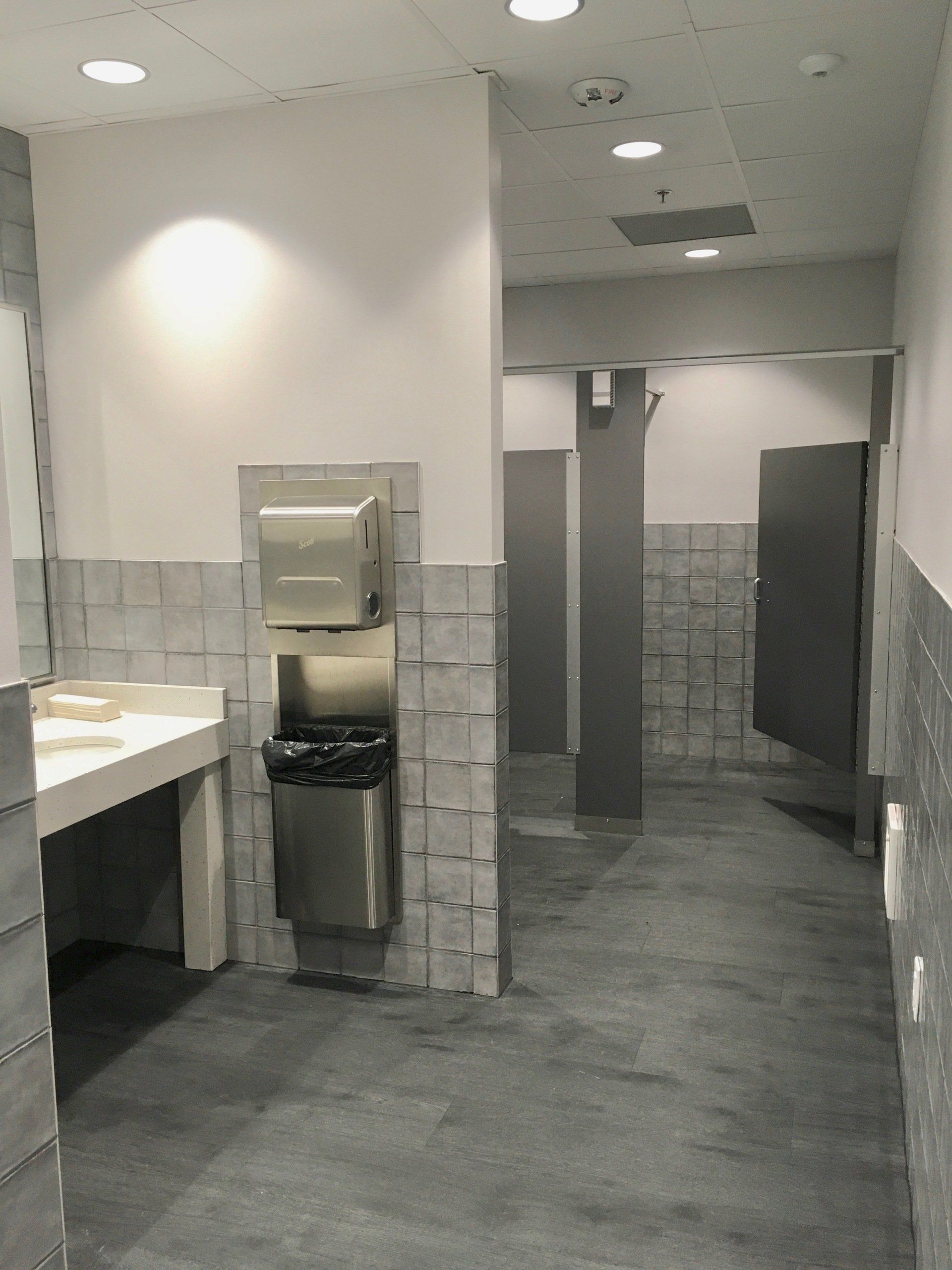 Public restroom with stalls, sink, paper towel dispenser, and trash can. Gray and white color scheme.