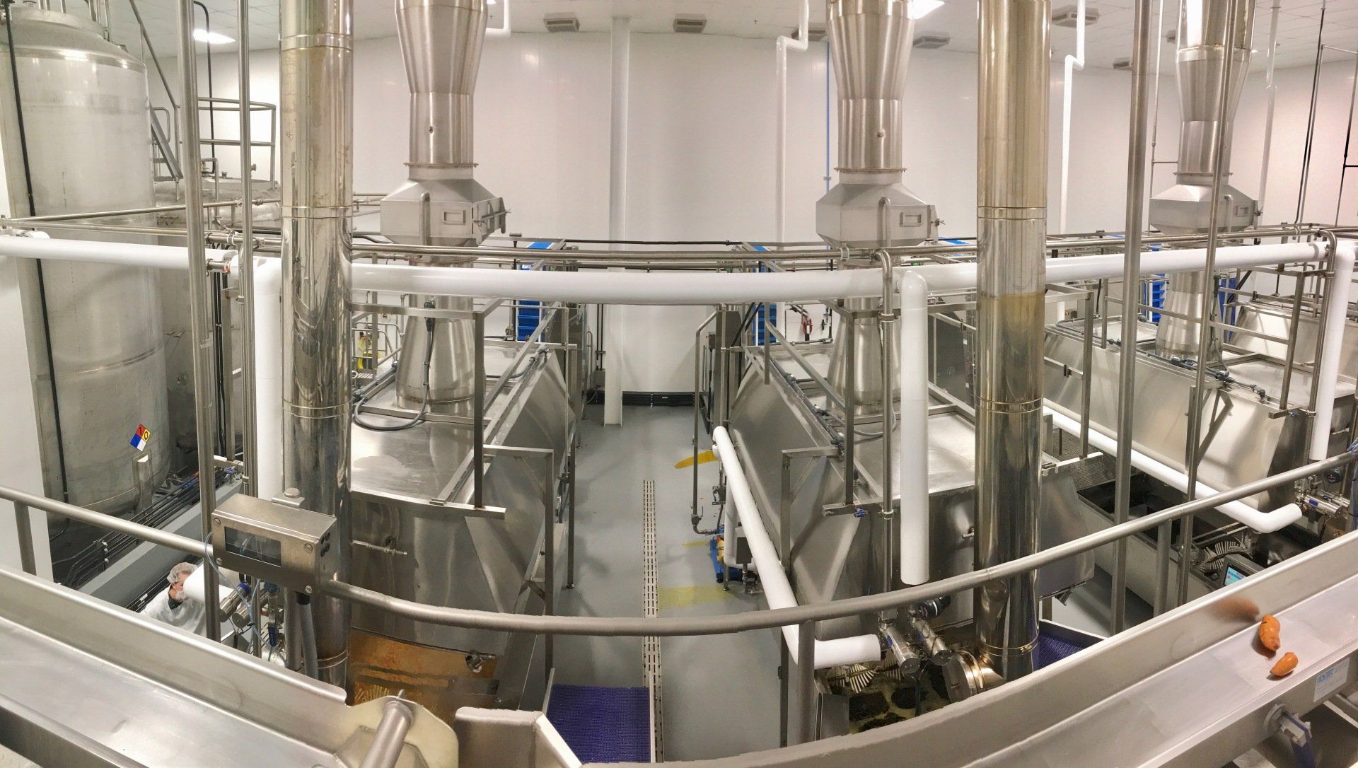 Industrial food processing equipment in a clean, white-walled factory setting. Stainless steel tanks, pipes, and conveyor belts.