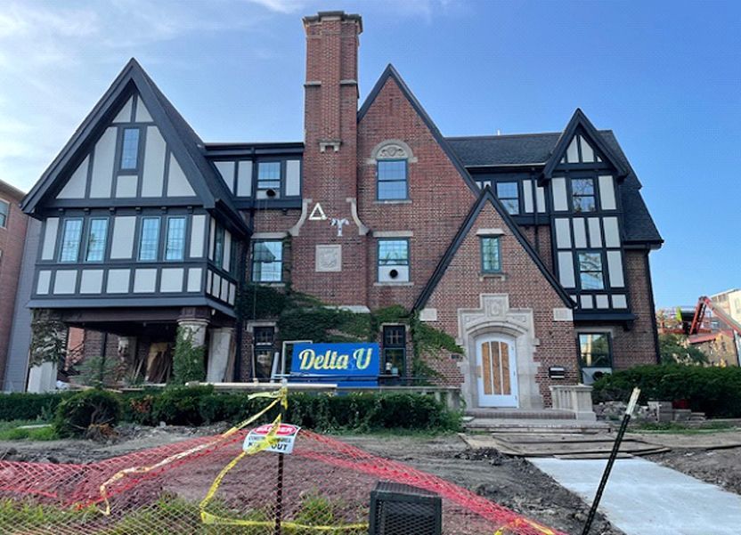 Tudor-style brick building with Delta U sign, under construction. Construction fencing surrounds the front yard.