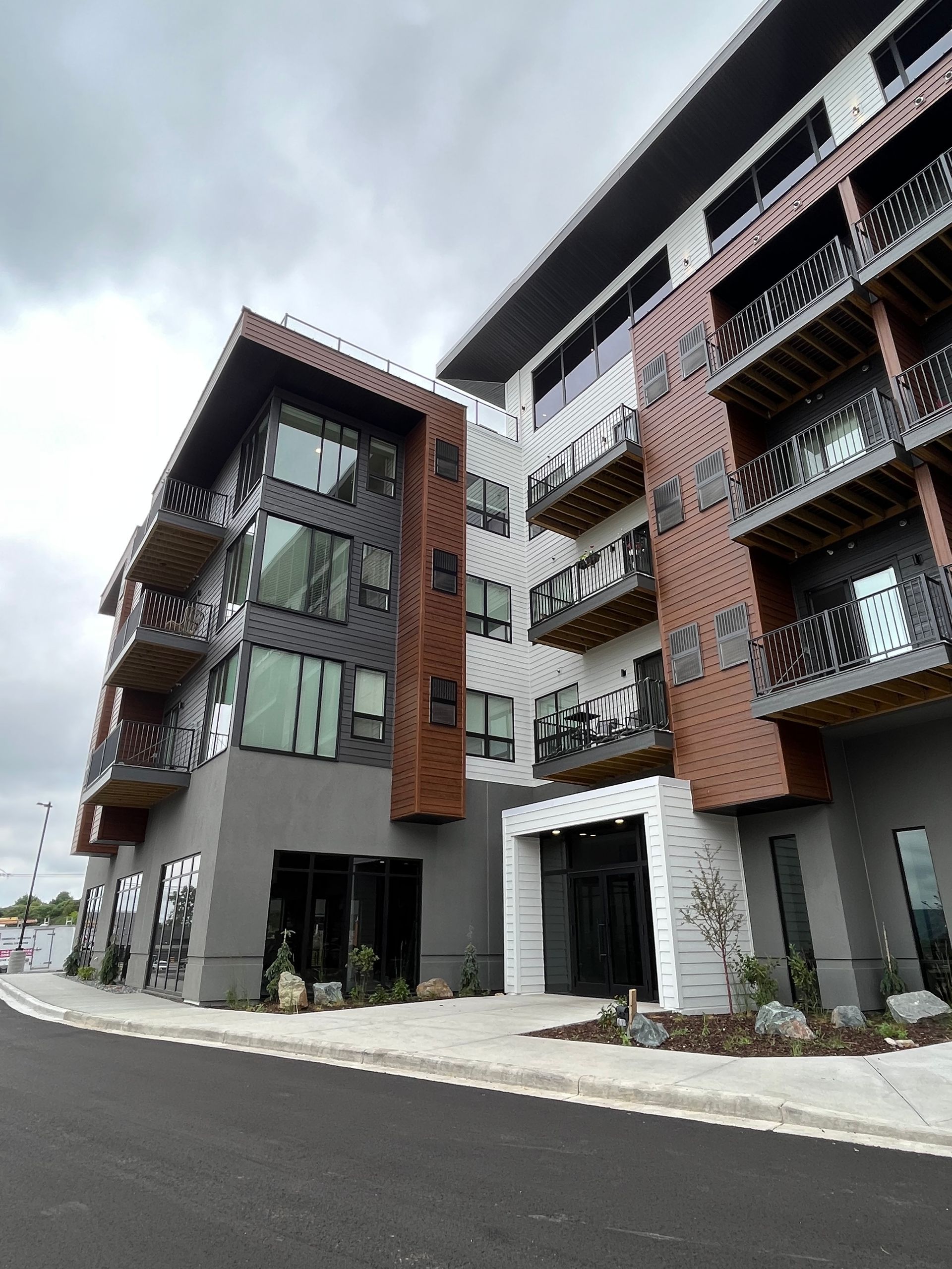 Modern multi-story apartment building with balconies, gray and brown facade, cloudy sky.