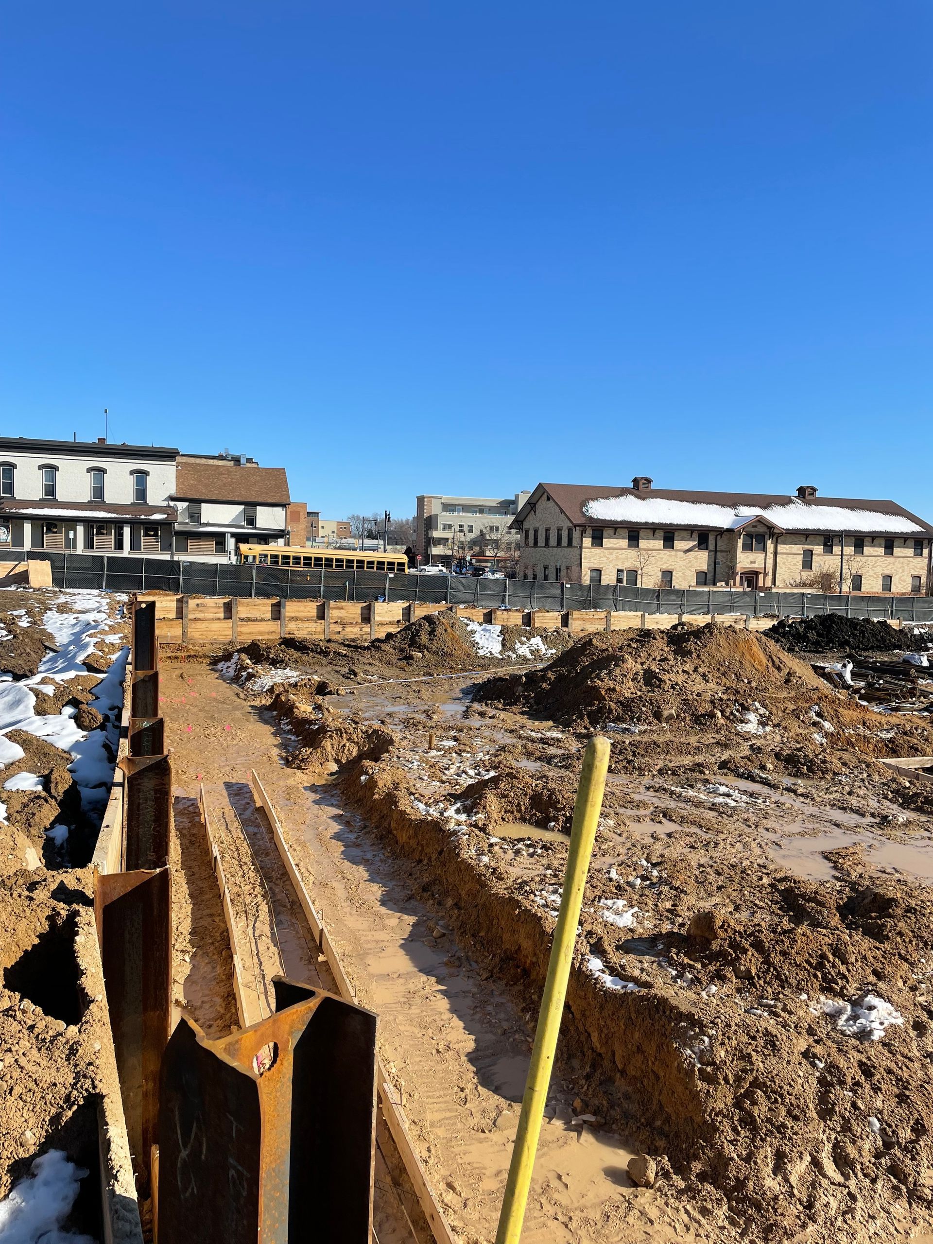 Construction site with dirt, metal barriers, and buildings under a bright blue sky.
