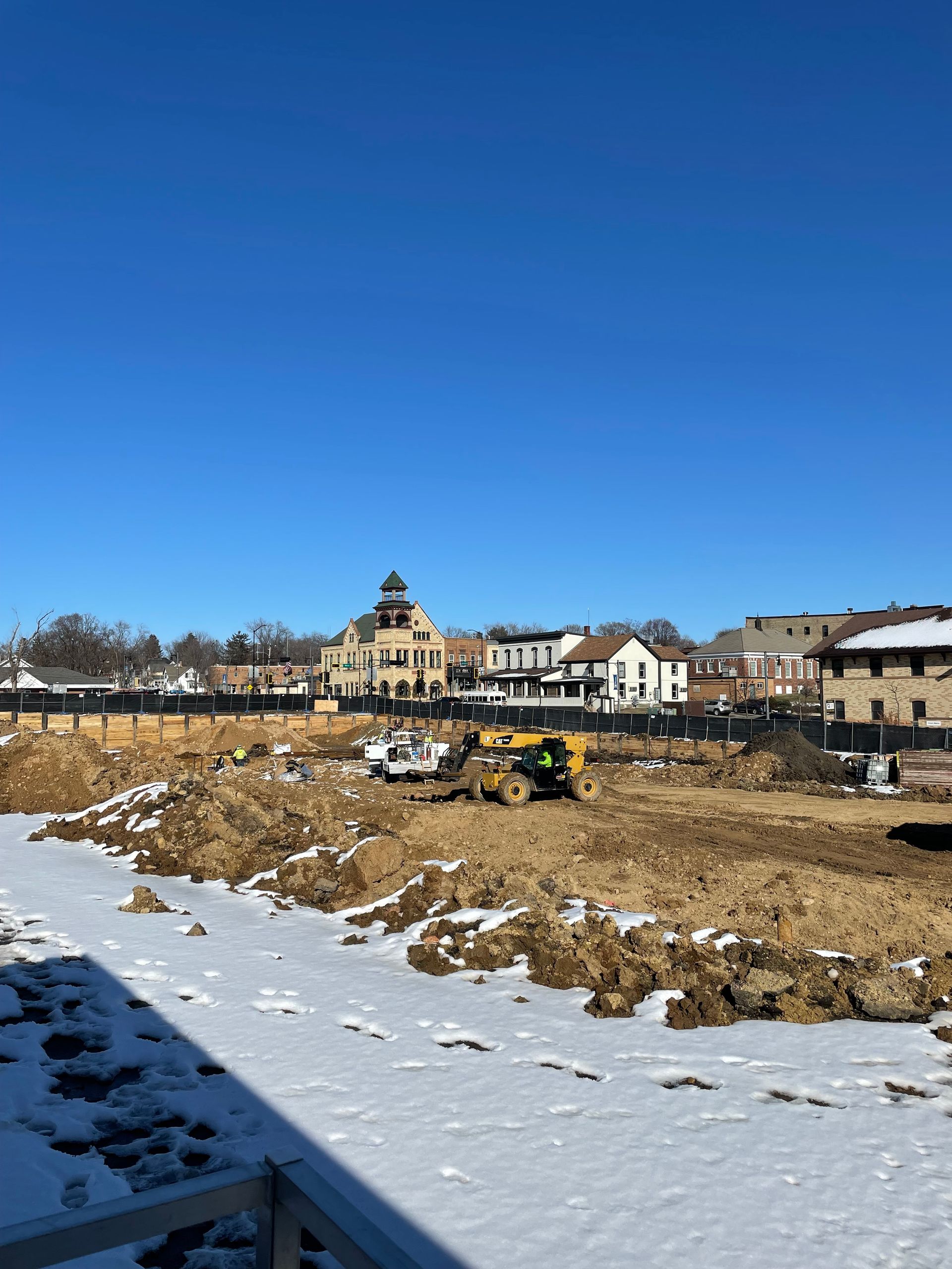 Construction site with earthmovers in foreground, buildings under a clear blue sky in the background. Snowy ground.