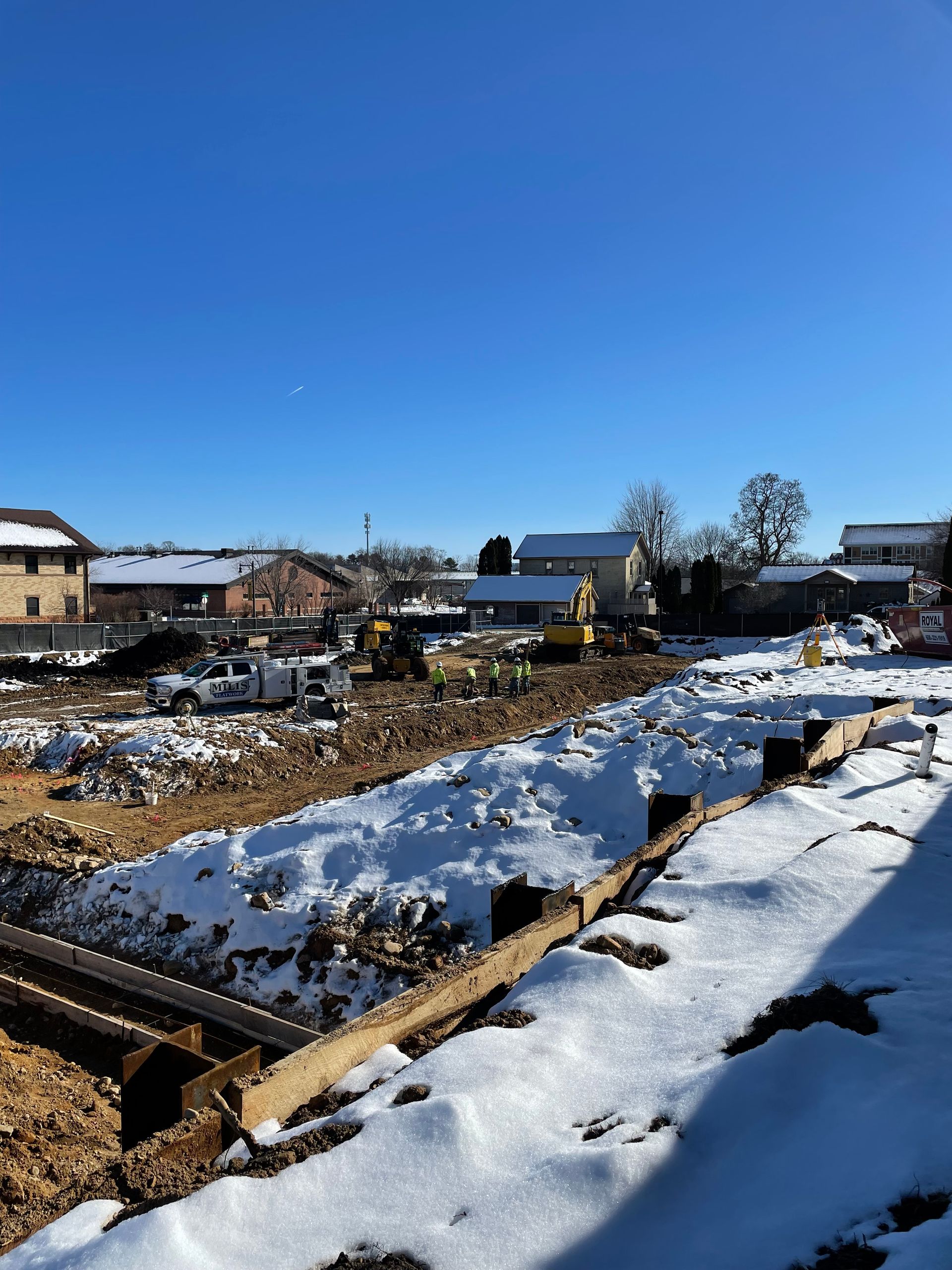 Construction site covered in snow, with buildings and equipment in the background under a clear blue sky.