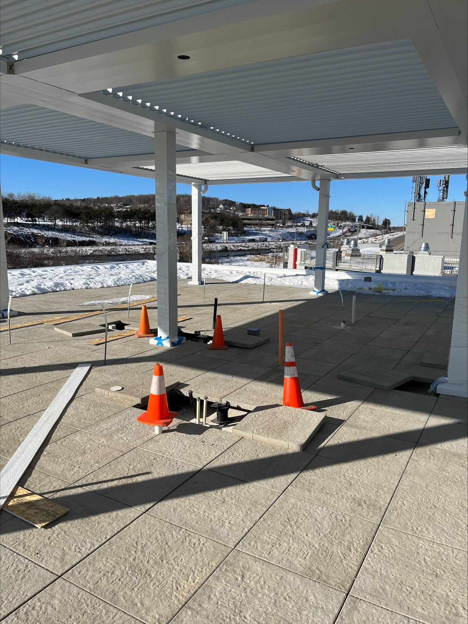 Construction site with orange cones on a tiled patio under a white pergola, overlooking a snowy landscape.