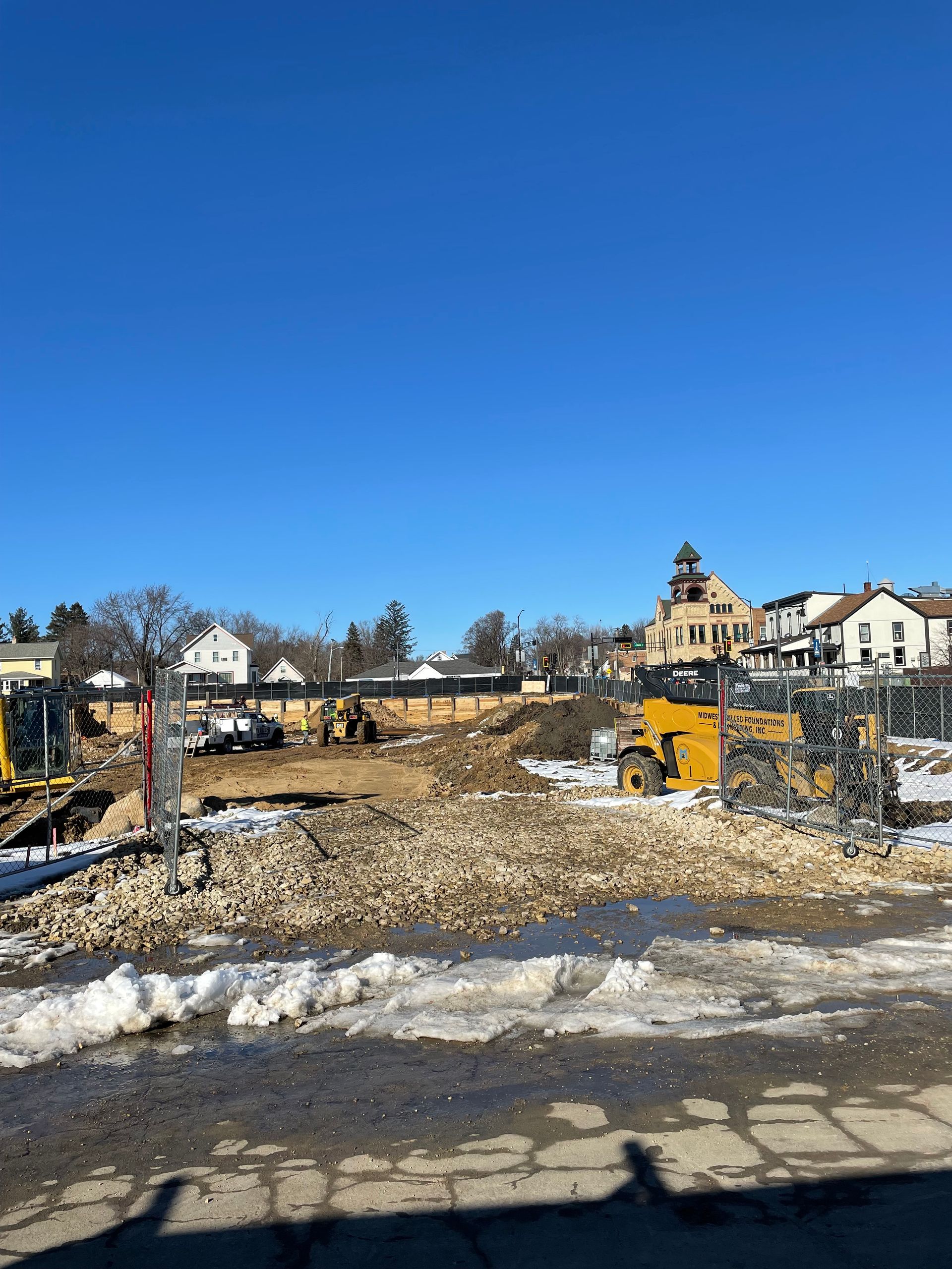 Construction site with rubble, excavators, and buildings under a blue sky.