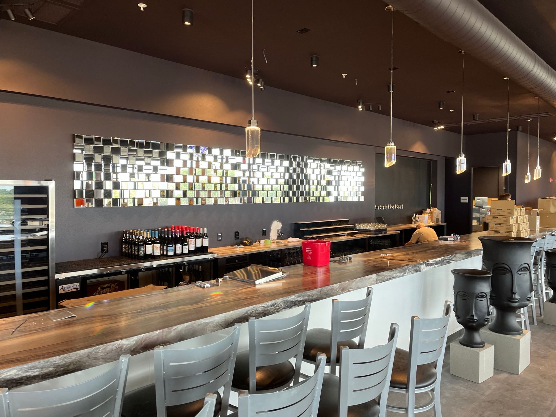 Bar interior with counter seating, wine bottles, and pendant lights. A mirrored wall adds a reflective pattern.