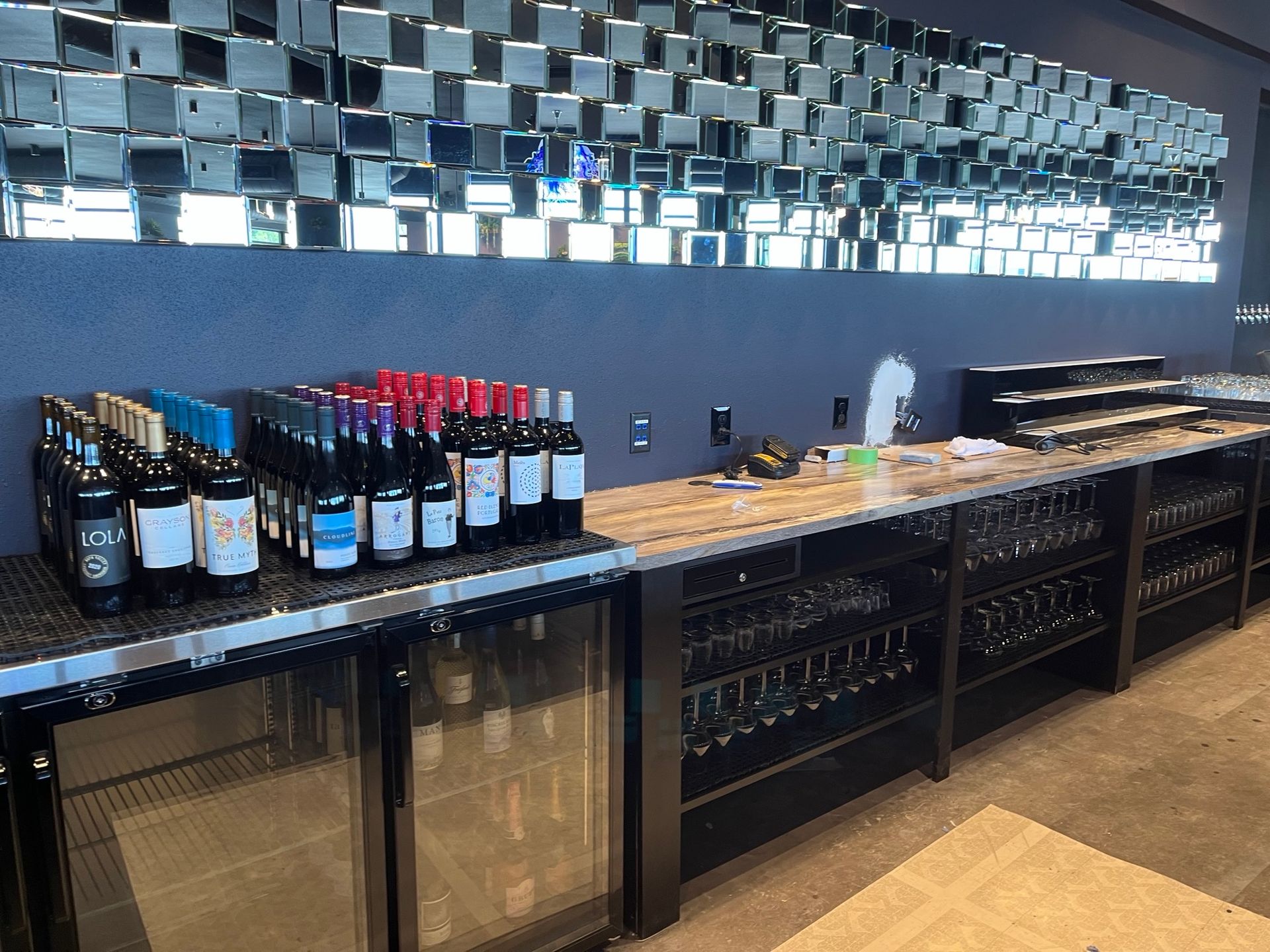 Wine bottles on a bar with mirrored tile wall.  Wine glasses hang below, cooler visible.