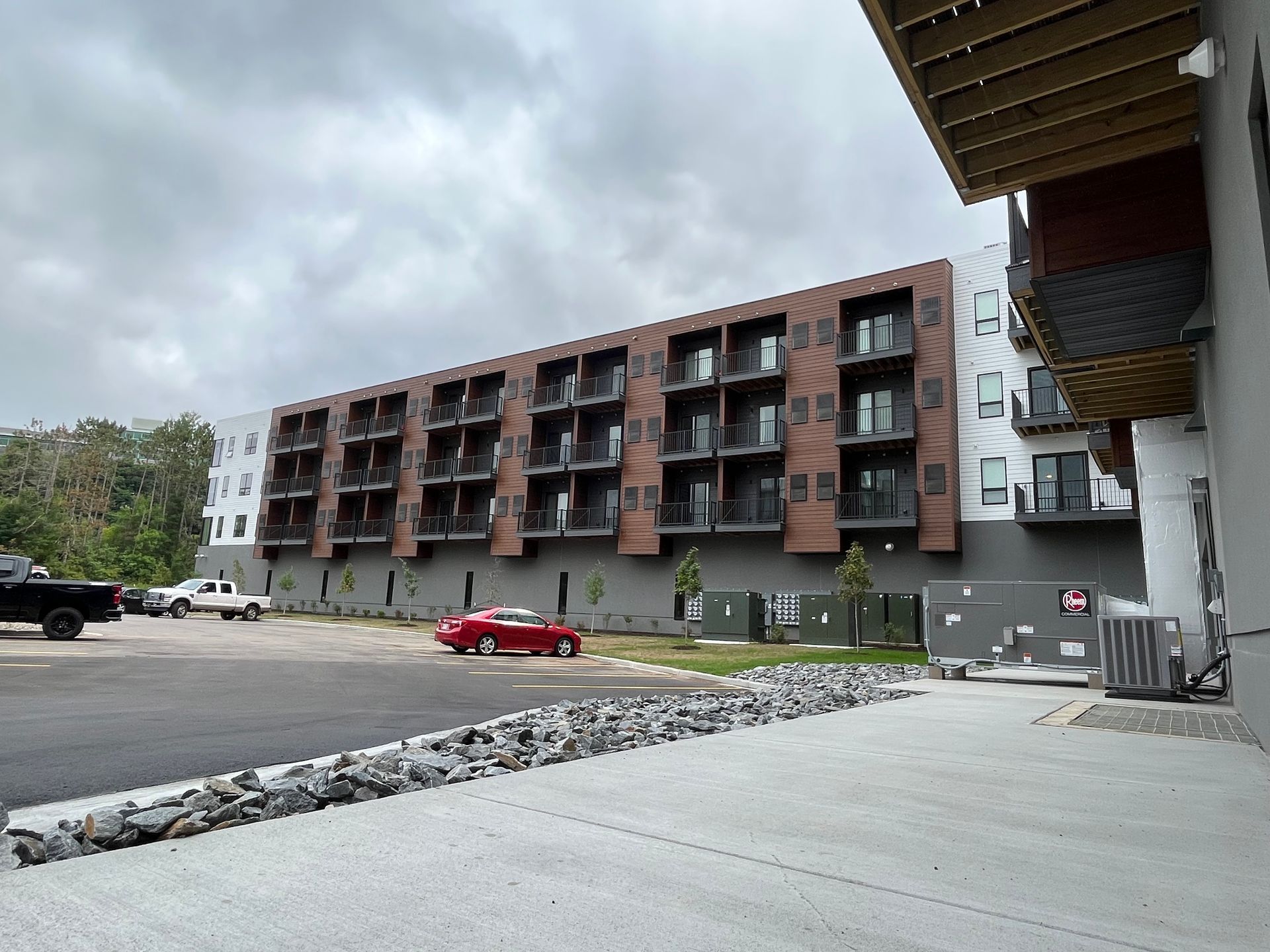 Apartment building with balconies and parking lot. Red car parked near building. Overcast sky.