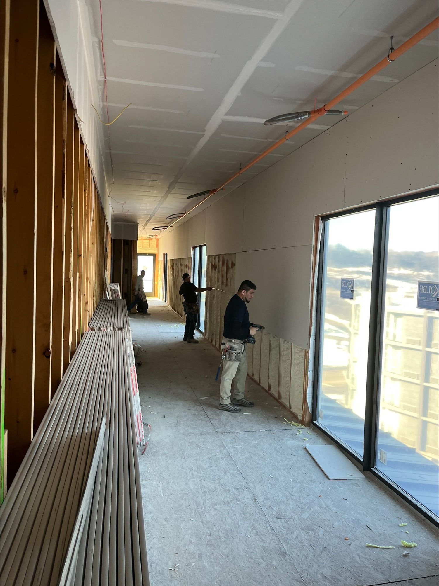 Construction workers inside a hallway installing drywall and insulation. Tall windows line one side.