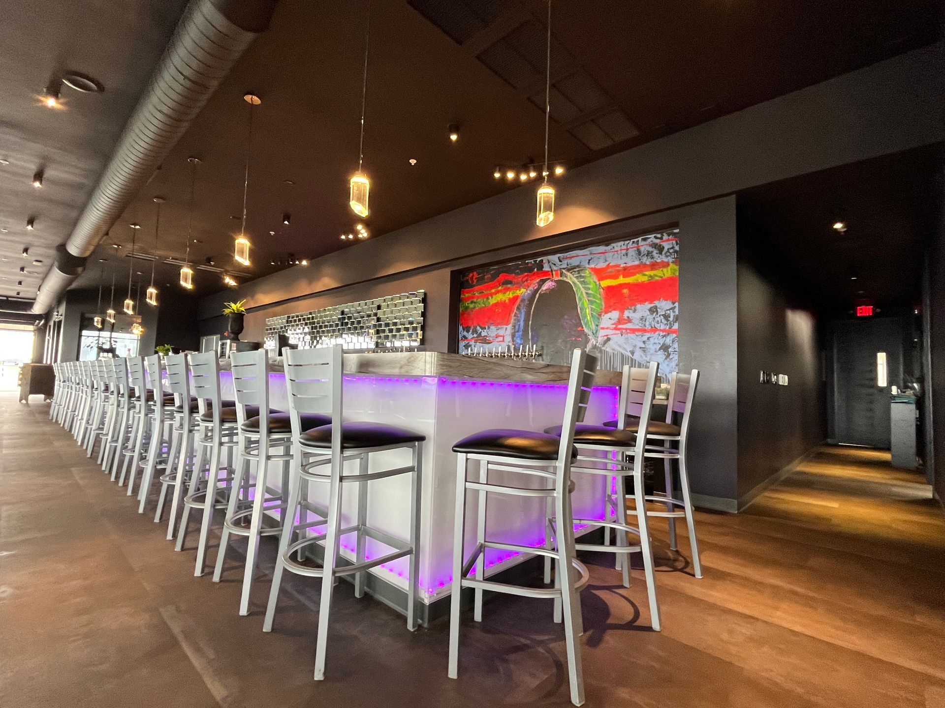 A long, lit bar with silver stools, purple underlighting, and a colorful abstract art piece in a dimly lit restaurant.