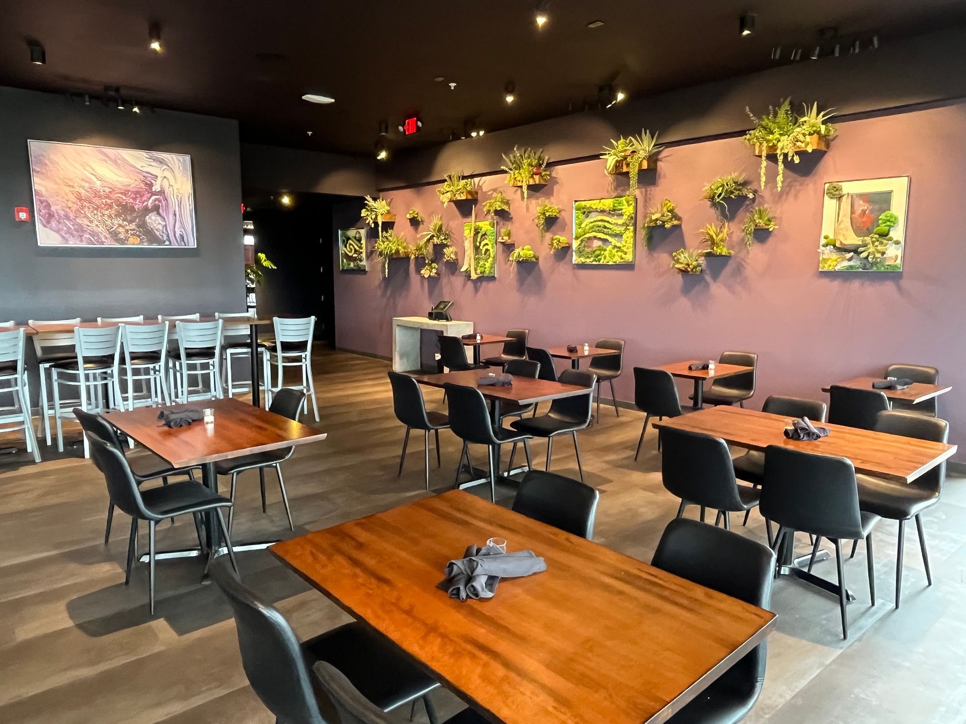 Restaurant interior with tables, chairs, bar seating, artwork, and wall plants.