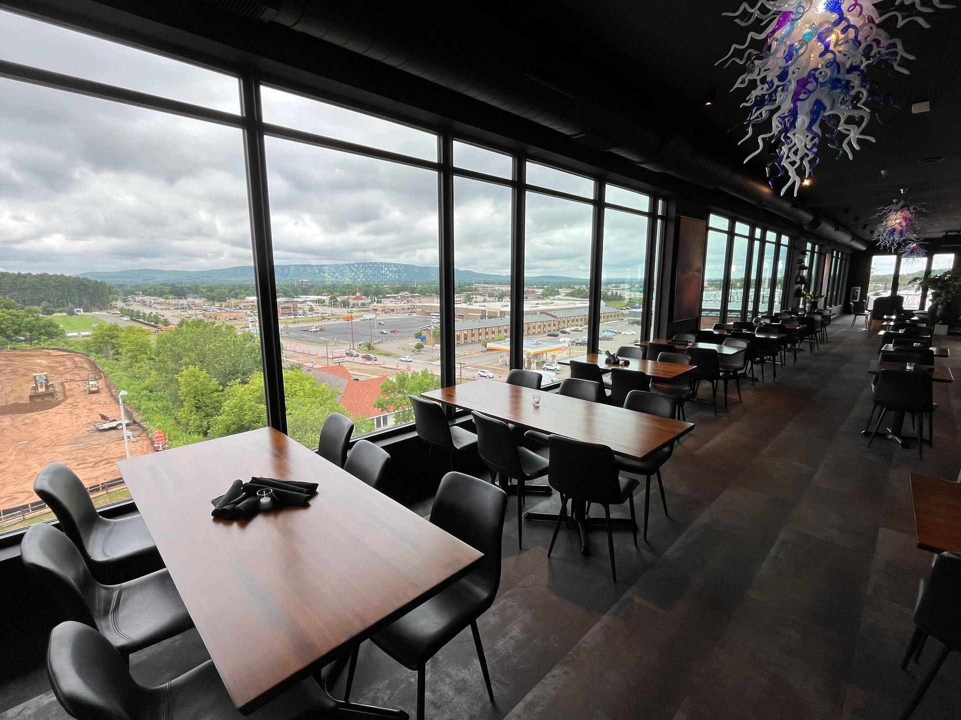 Restaurant interior with cityscape view through large windows. Tables and black chairs arranged for dining.