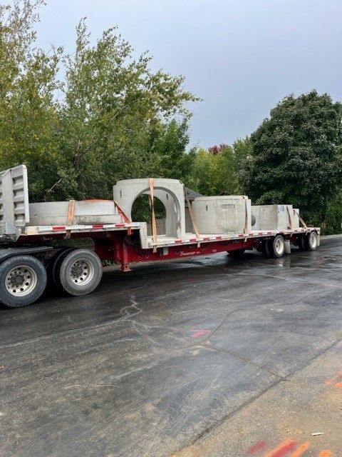 Flatbed truck carrying large, concrete culvert sections on a wet, gray surface. Trees in the background.